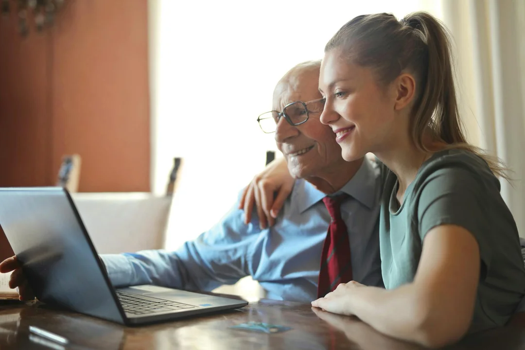 A senior man with glasses and a younger woman sit at a table looking at a laptop, discussing HOA rule changes.