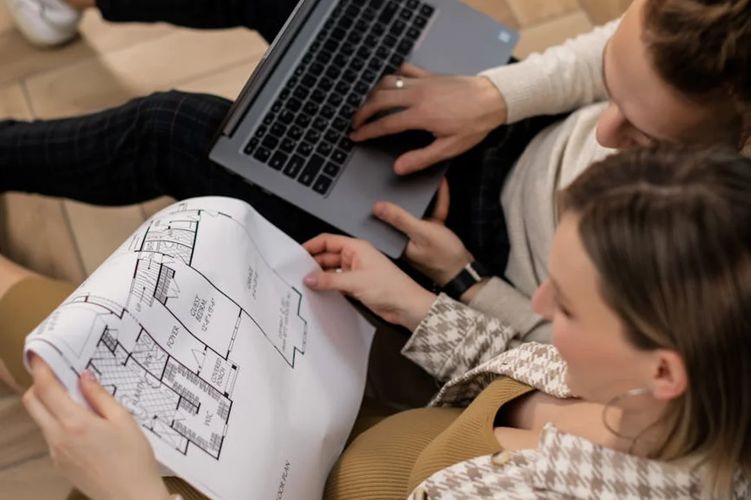 Two people review architectural blueprints with a laptop nearby