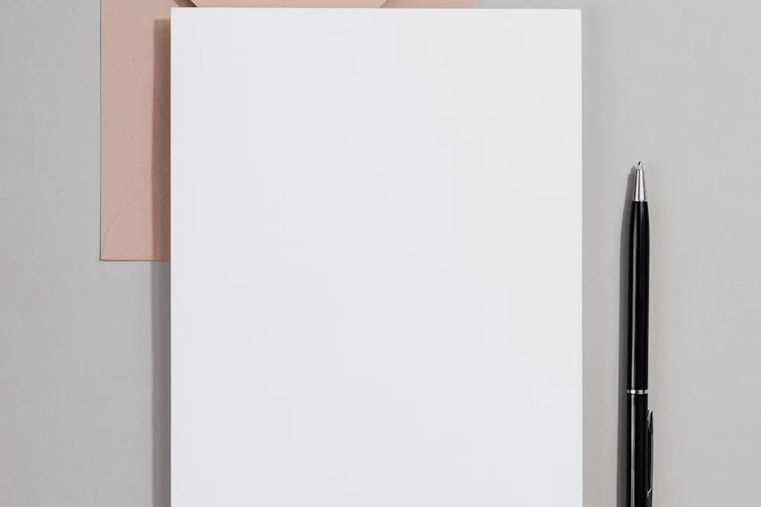 A blank white notebook on a light gray surface with a black pen to the right and a pale pink card peeking from behind, symbolizing planning and documentation for a resignation and handover.