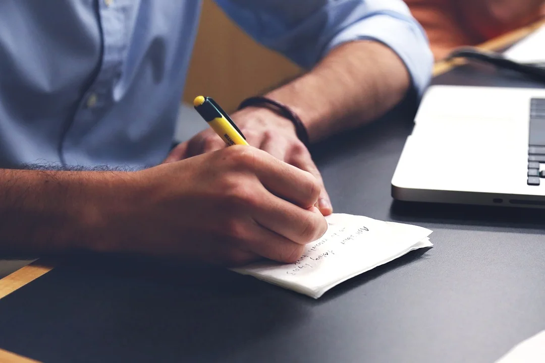 Person writing on a small notepad with a yellow pencil, laptop nearby, planning an HOA software upgrade.