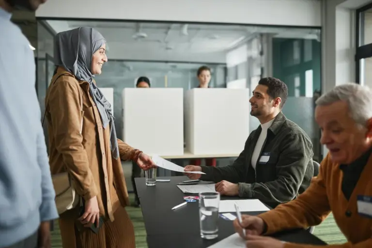A woman wearing a hijab hands a document to a man seated at a conference table during an HOA meeting, with other participants visible in the background.
