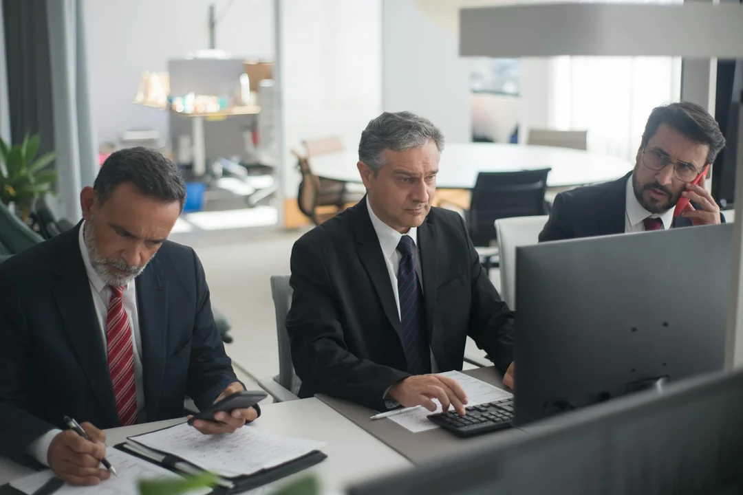 Three professionals in business attire review documents and a computer screen in a modern office to plan an HOA reserve fund strategy.