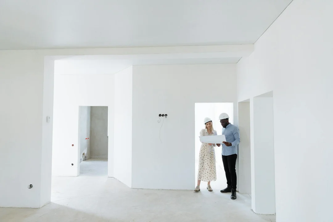 Two professionals in hard hats examine blueprints in a bright, empty construction space