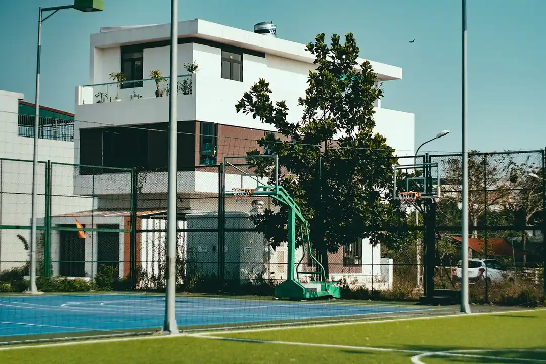 Fenced outdoor basketball court with a green hoop in front of a modern white building and trees under a clear blue sky.