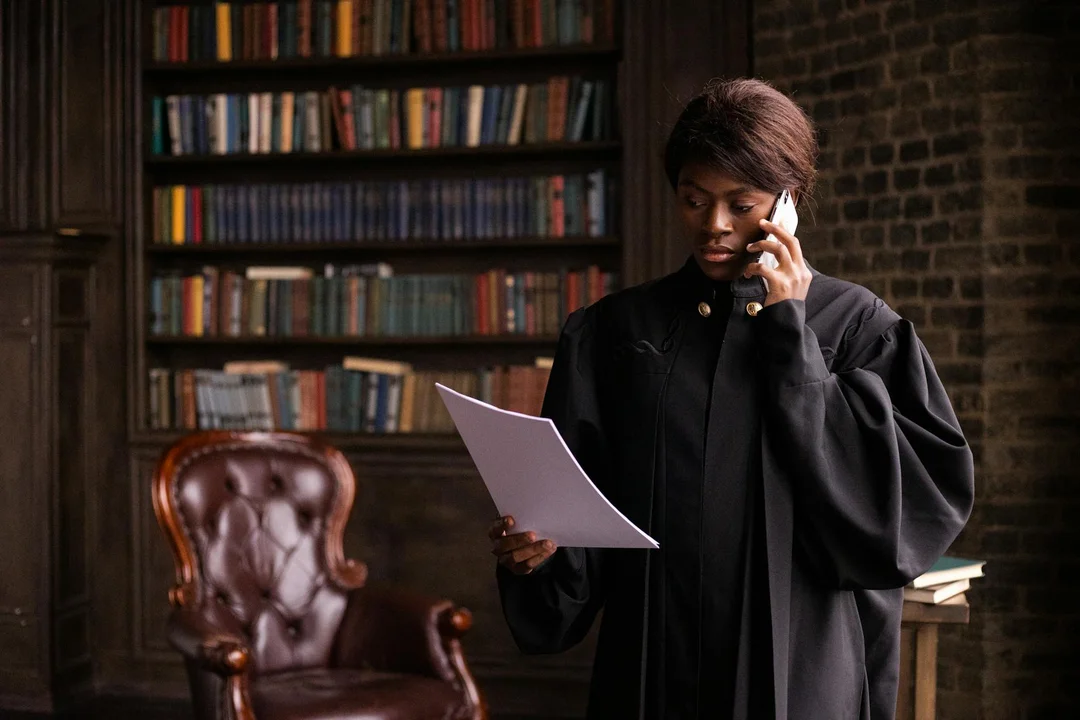 A legal professional wearing a black robe stands in a library, holding a document and speaking on a cellphone.