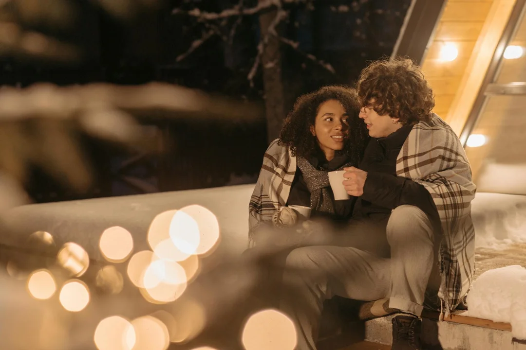 Couple sitting outdoors on a snowy doorstep wrapped in a blanket, surrounded by warm holiday lights.
