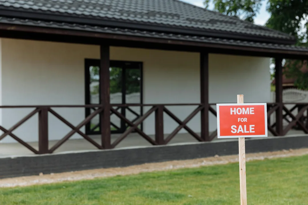 A house with a red 'Home for Sale' sign in the front yard and a wooden porch railing.