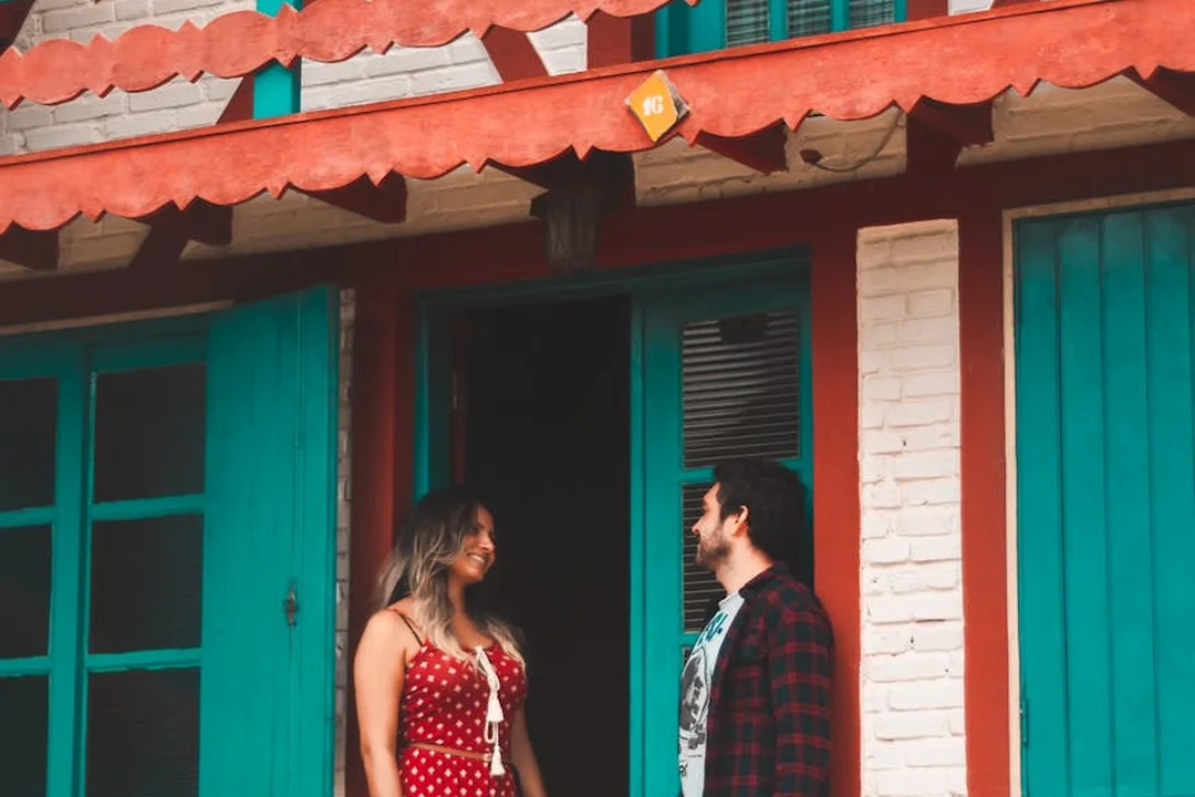 A couple stands outside a colorful house with turquoise doors and red trim, smiling at each other.