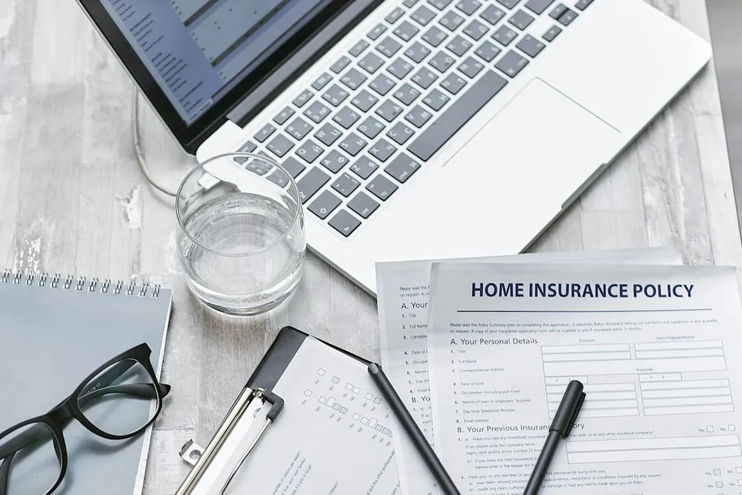 Desk scene with a laptop, a glass of water, home insurance policy form, notebook, clipboard, glasses, and a pen.