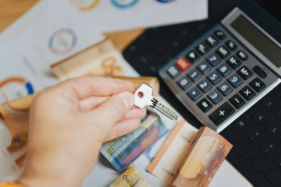 Close-up of a hand holding a key above scattered euro banknotes and a calculator, symbolizing homeownership decisions and financial planning.