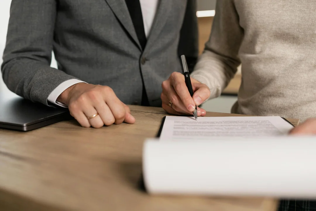 Two people signing documents at a wooden desk in a professional setting, with a laptop nearby.