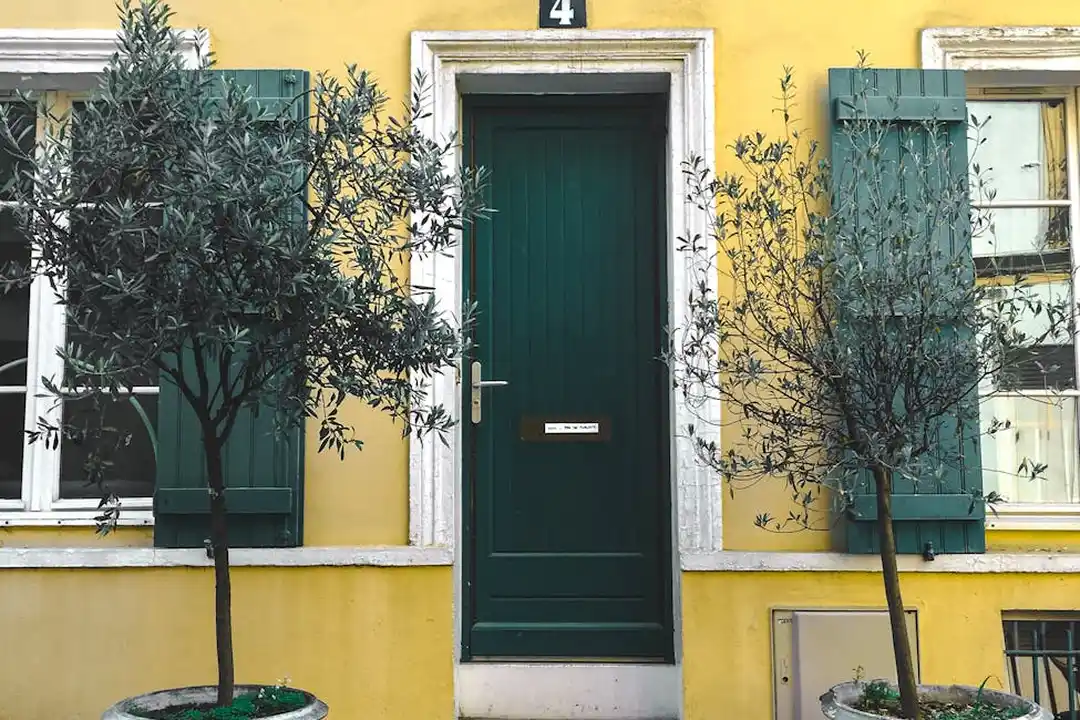 Front view of a yellow house with a green door and matching green window shutters, with small trees planted along the front.