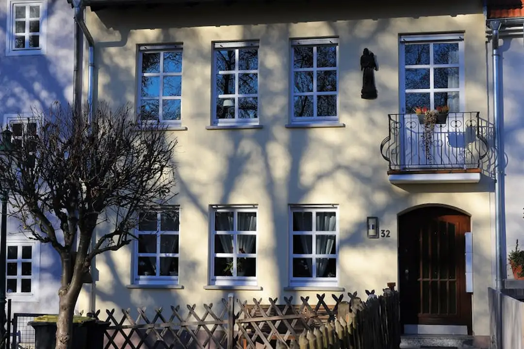 Front view of a two-story house with multiple white-trimmed windows, a small balcony with a wrought-iron railing, a wooden front door, a tree, and a low wooden fence.
