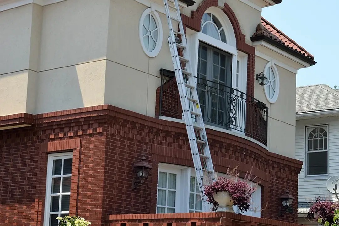 Two-story suburban house with a tall ladder leaning against the balcony, brick lower facade and beige upper walls.