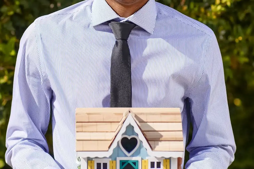 A person in a light purple striped shirt and dark tie holds a small wooden model house, illustrating lenders evaluating HOA fees during mortgage approval.