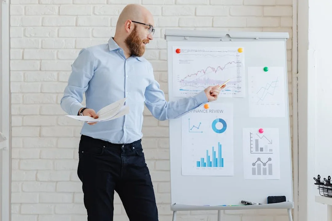 Bearded man in a light-blue shirt presenting financial charts on a whiteboard in an office, illustrating HOA budgeting and fee allocation.