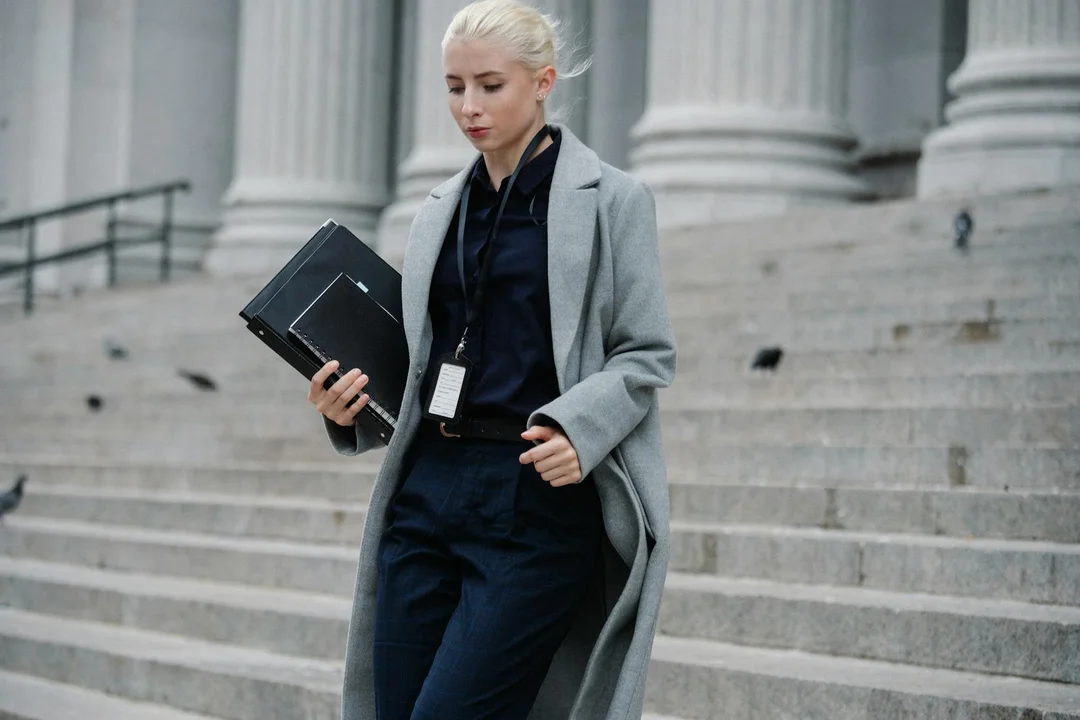 Professional woman in a gray coat carrying a binder and documents walks down stone steps outside a stately building with columns.