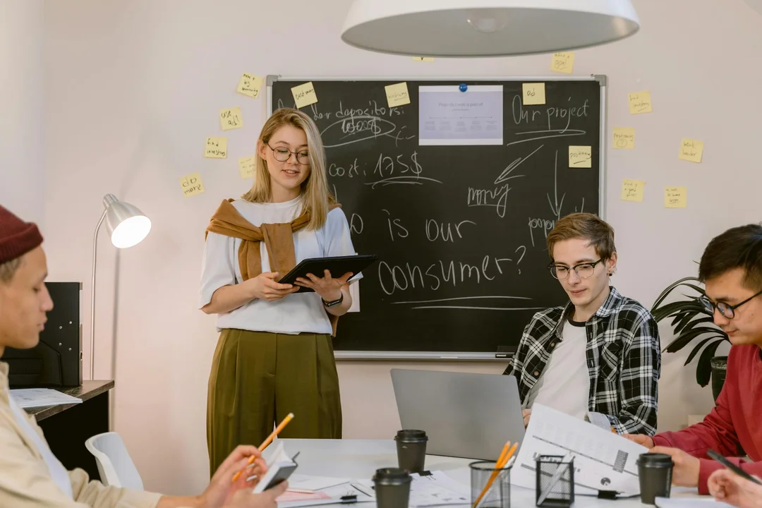 A diverse group of people around a conference table collaborating, with a woman standing and taking notes as a chalkboard behind them is filled with sticky notes and diagrams.
