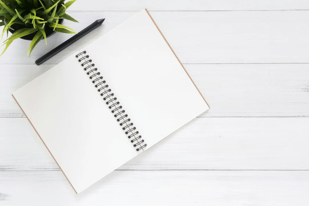 Open notebook with a spiral binding, pen, and a small plant on a white wooden desk, representing reviewing HOA financial documents.