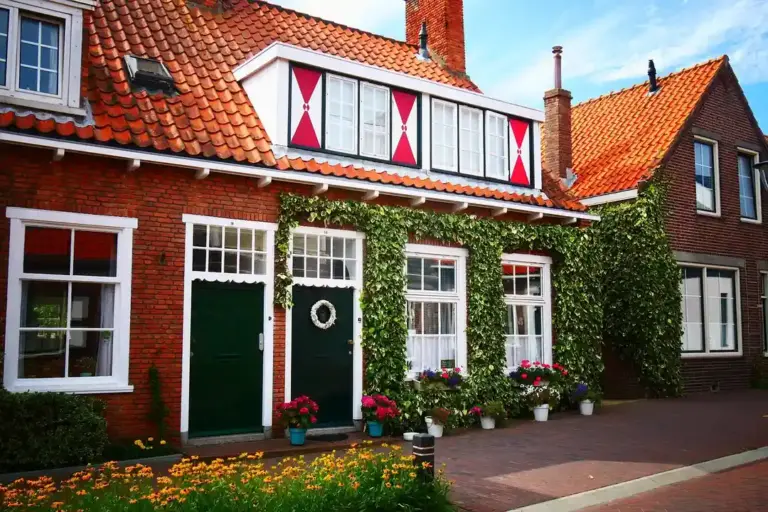 Row of brick houses with white-framed windows and ivy climbing the walls, red-tiled roofs, and potted flowers along a sidewalk.