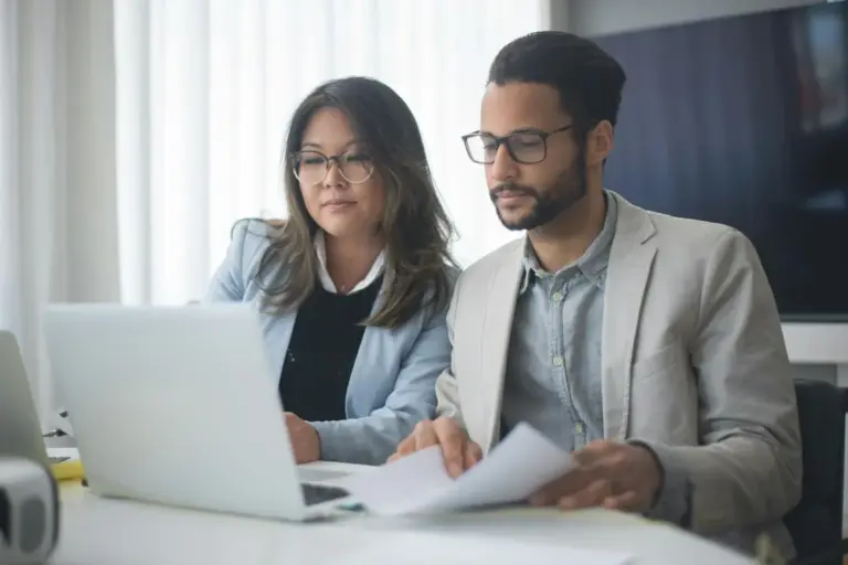 Two professionals sit at a desk with a laptop and papers, reviewing HOA governing documents in a bright office