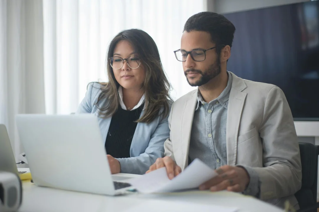 Two professionals sit at a desk with a laptop and papers, reviewing HOA governing documents in a bright office