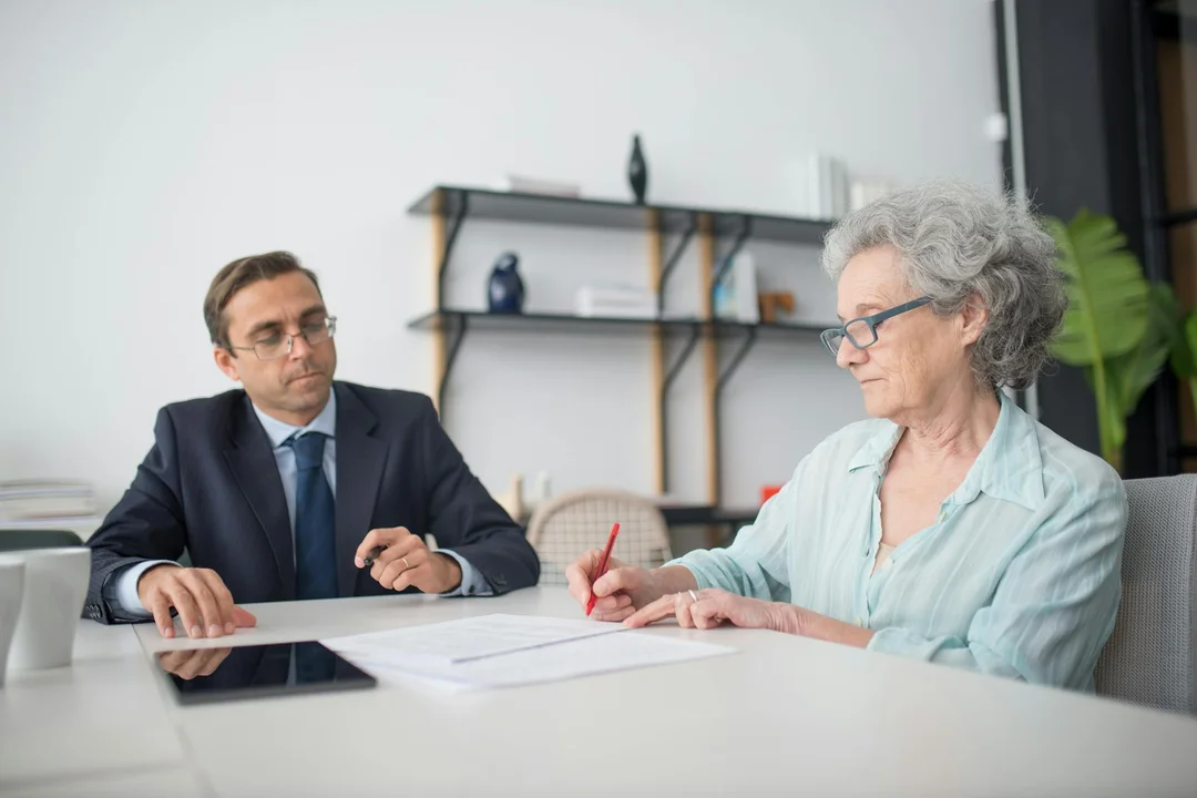 Two professionals at a table in a bright office, a man in a suit and an older woman with glasses reviewing documents.