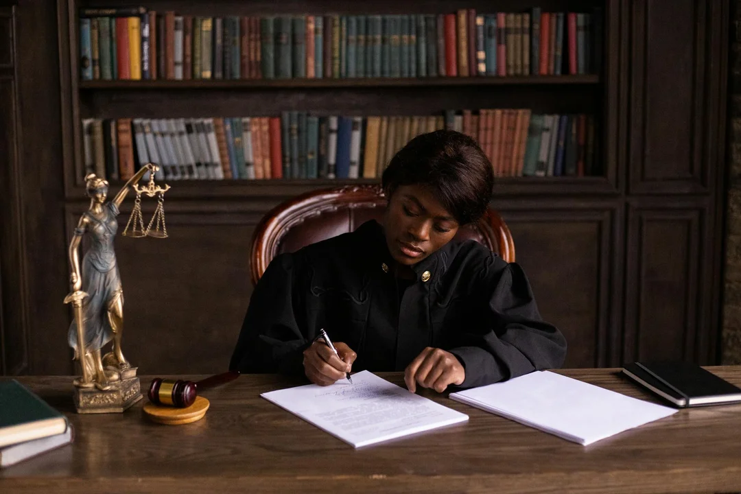 Judge at a wooden desk signing papers, with a scales of justice statue and books in the background.