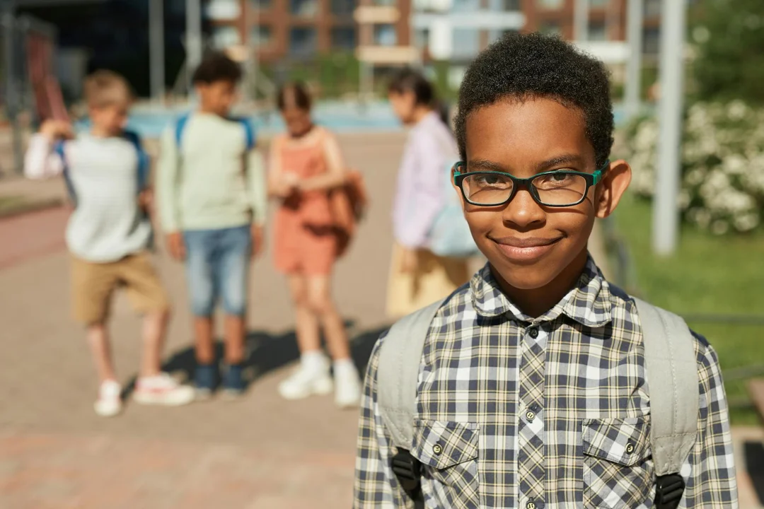 A smiling young boy with glasses and backpack stands in the foreground, with a group of children at a park in the background, illustrating community planning for a new amenity.