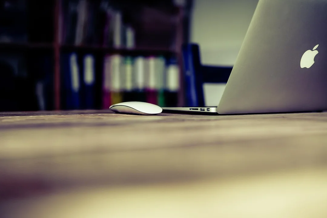 Close-up of a laptop and mouse on a wooden desk with a blurred book-filled background.