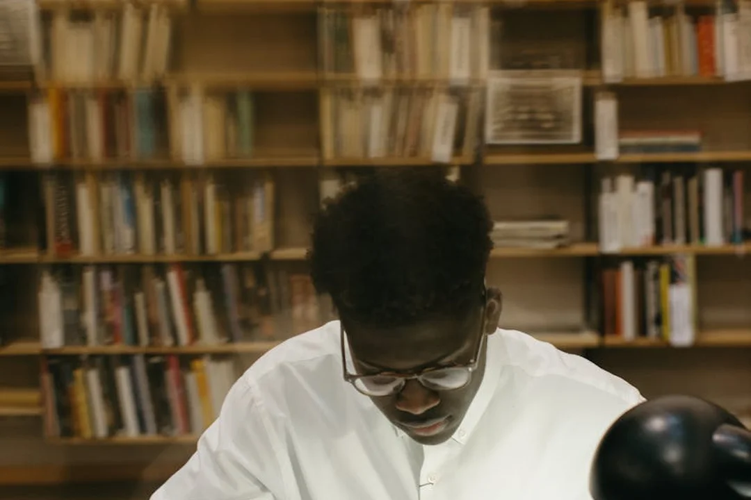 Person wearing glasses and a white shirt sits at a table in a library, studying with bookshelves in the background.