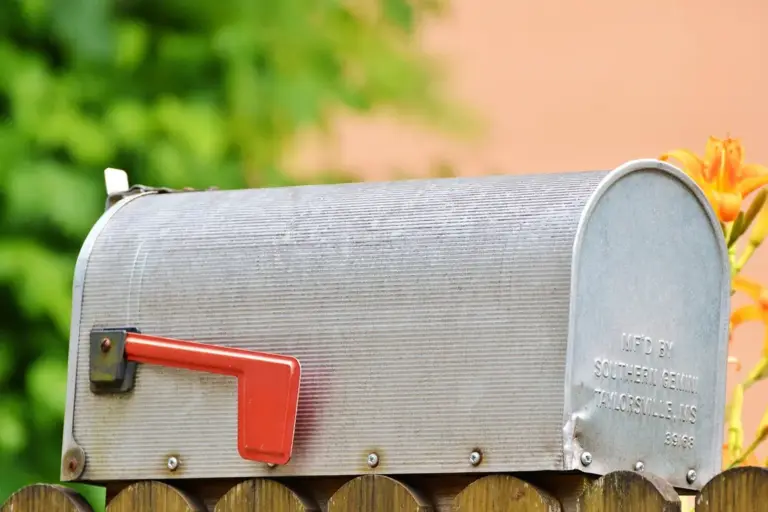 Metal mailbox with a red flag raised, mounted on a wooden fence with blurred greenery in the background.