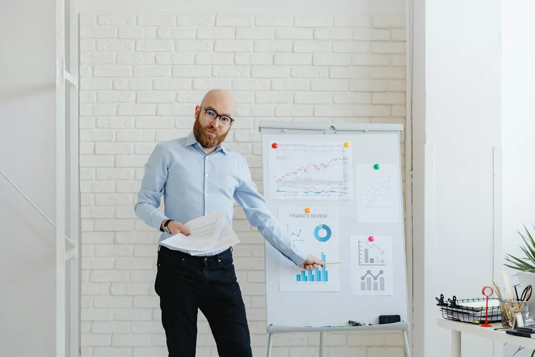 Bearded man in a light blue shirt presenting charts on a whiteboard in a bright office.