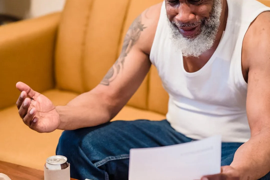 An older man with a gray beard wearing a white sleeveless shirt sits on a yellow couch, looking down at a document on a coffee table.