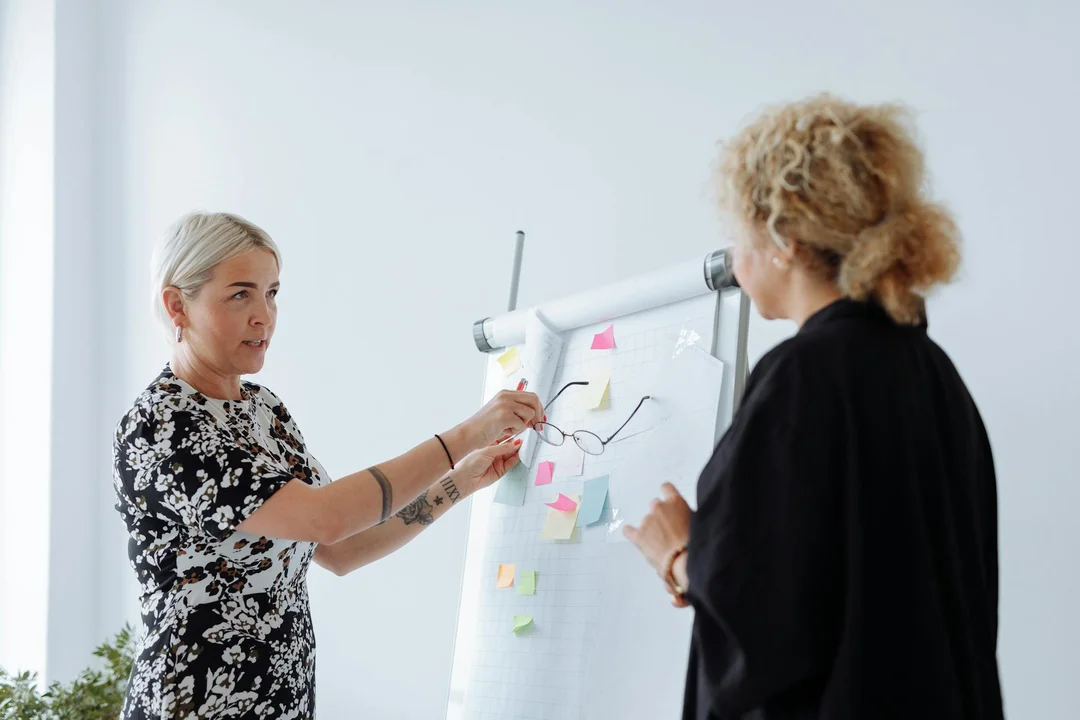 Two women in a professional setting stand near a flip chart with sticky notes, discussing mediation options.