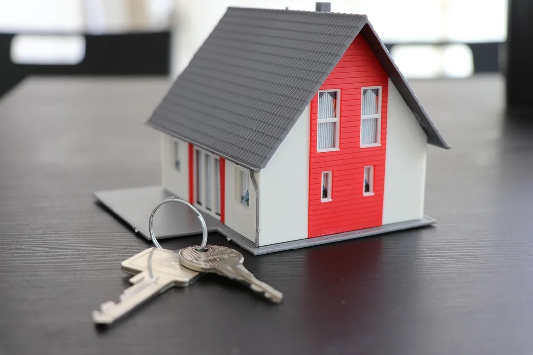 A miniature red-and-white house model with a gray roof sits on a dark surface next to a set of keys, illustrating escrow and HOA payment concepts.