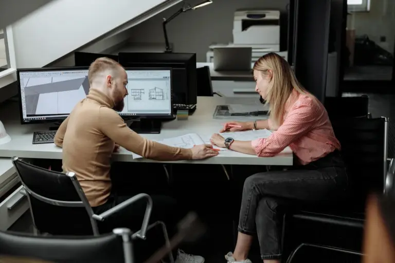 Two design professionals discuss architectural plans at a desk, with a computer displaying 3D blueprints