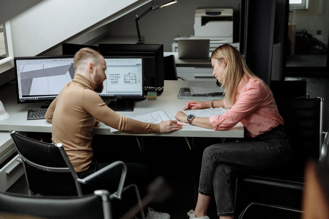 Two design professionals discuss architectural plans at a desk, with a computer displaying 3D blueprints