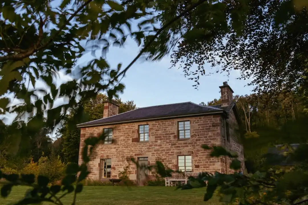 A brick two-story house with a dark roof, framed by green trees and shrubbery.