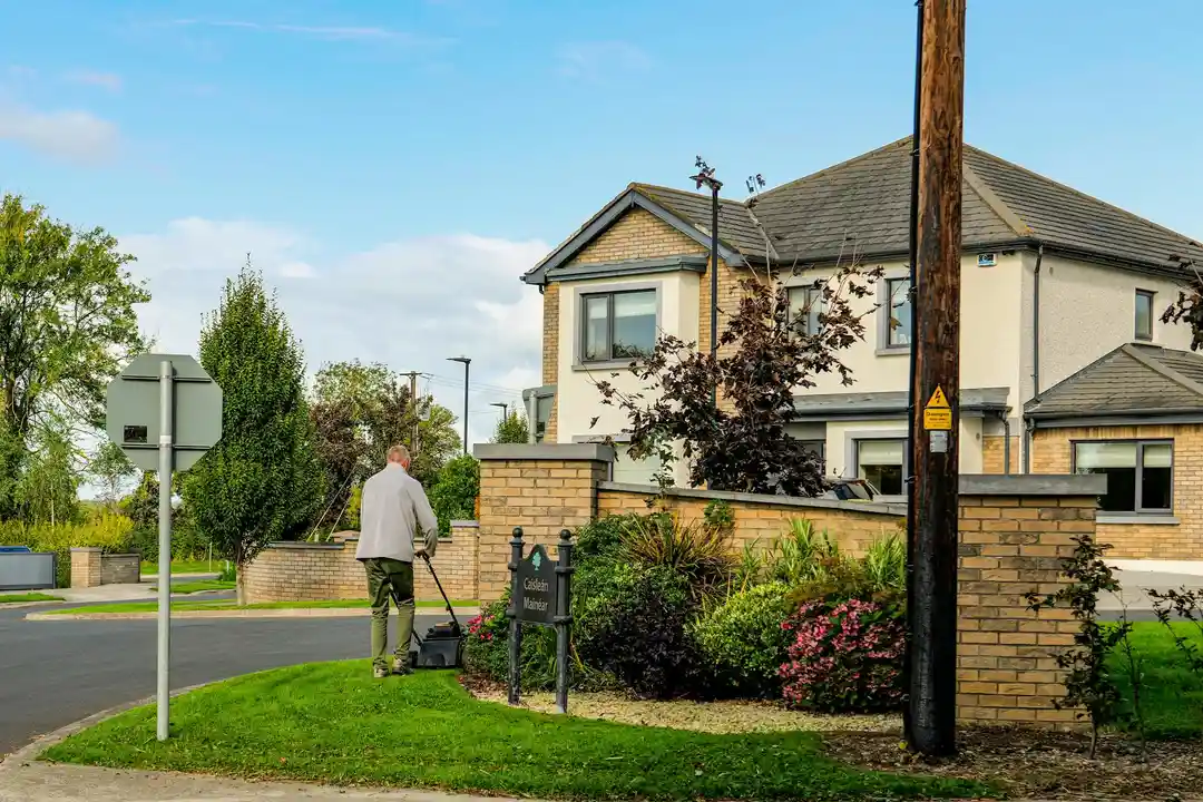 Person pushing a lawn mower to mow a front lawn beside a beige house with a brick wall and shrubs in a suburban neighborhood.
