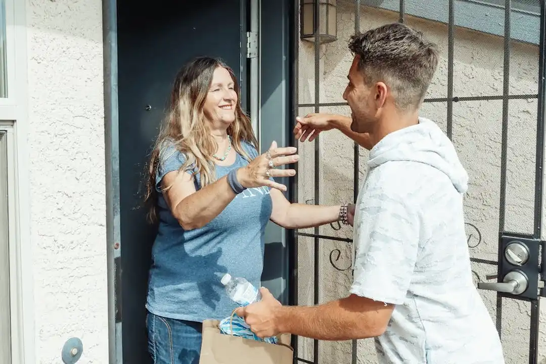 Two adults standing at the doorway of a home in an HOA community, smiling and talking as they greet each other near an iron gate.