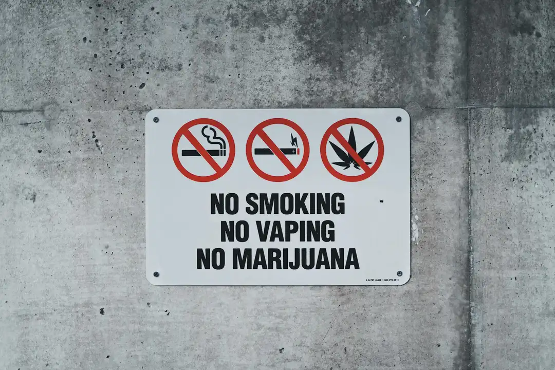 White rectangular sign on a gray concrete wall displaying three prohibition icons for smoking, vaping, and marijuana, with the text 'NO SMOKING NO VAPING NO MARIJUANA'.