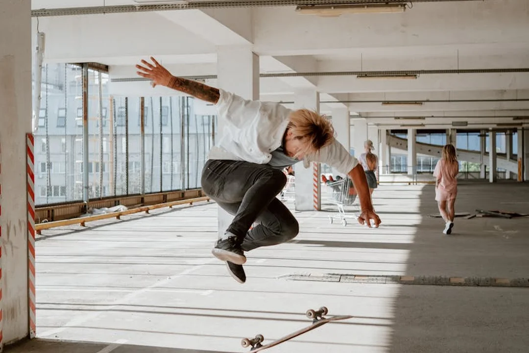 A person skateboarding and mid-air jump inside a spacious, sunlit indoor area with large windows; another person stands in the background.