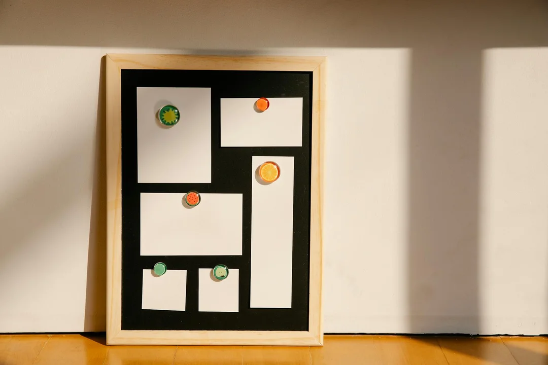 Framed display board with blank white papers and colorful circular stickers arranged on a wooden floor against a white wall.