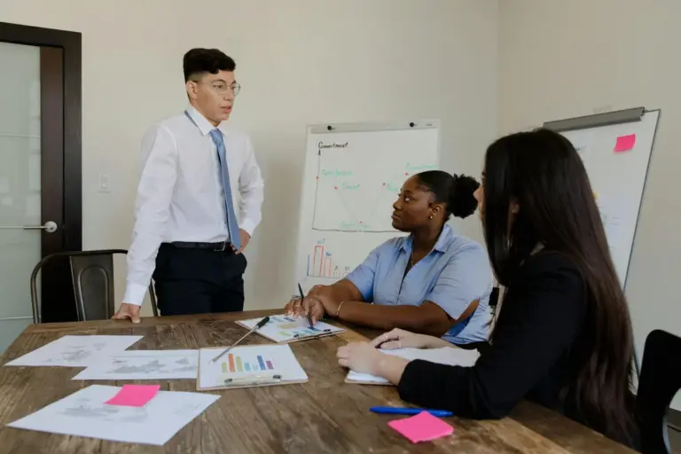 A business meeting in a conference room: a man wearing a white shirt and tie stands at the head of a table while two women sit and review documents, with charts on a whiteboard in the background.