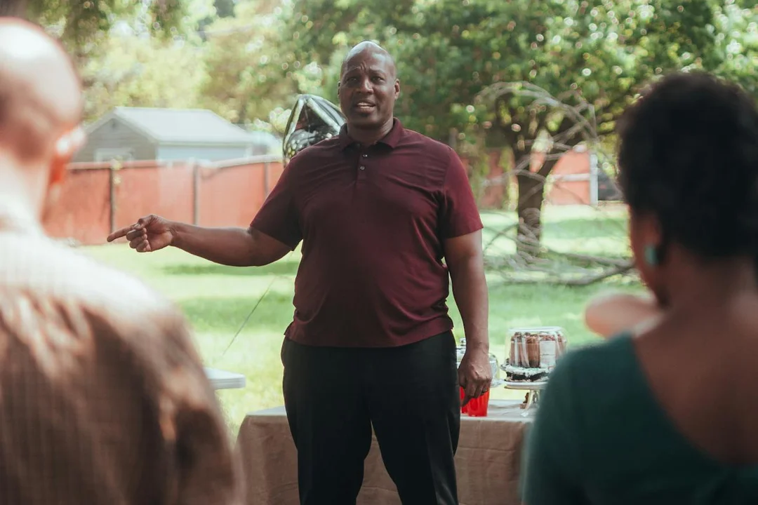 A community leader in a maroon polo speaks to neighbors outdoors during a HOA meeting, encouraging constructive conversation.