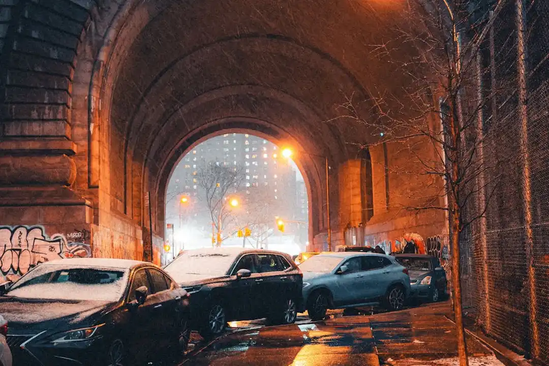 Cars parked along a dimly lit brick tunnel underpass with graffiti on the walls and orange streetlights casting a glow
