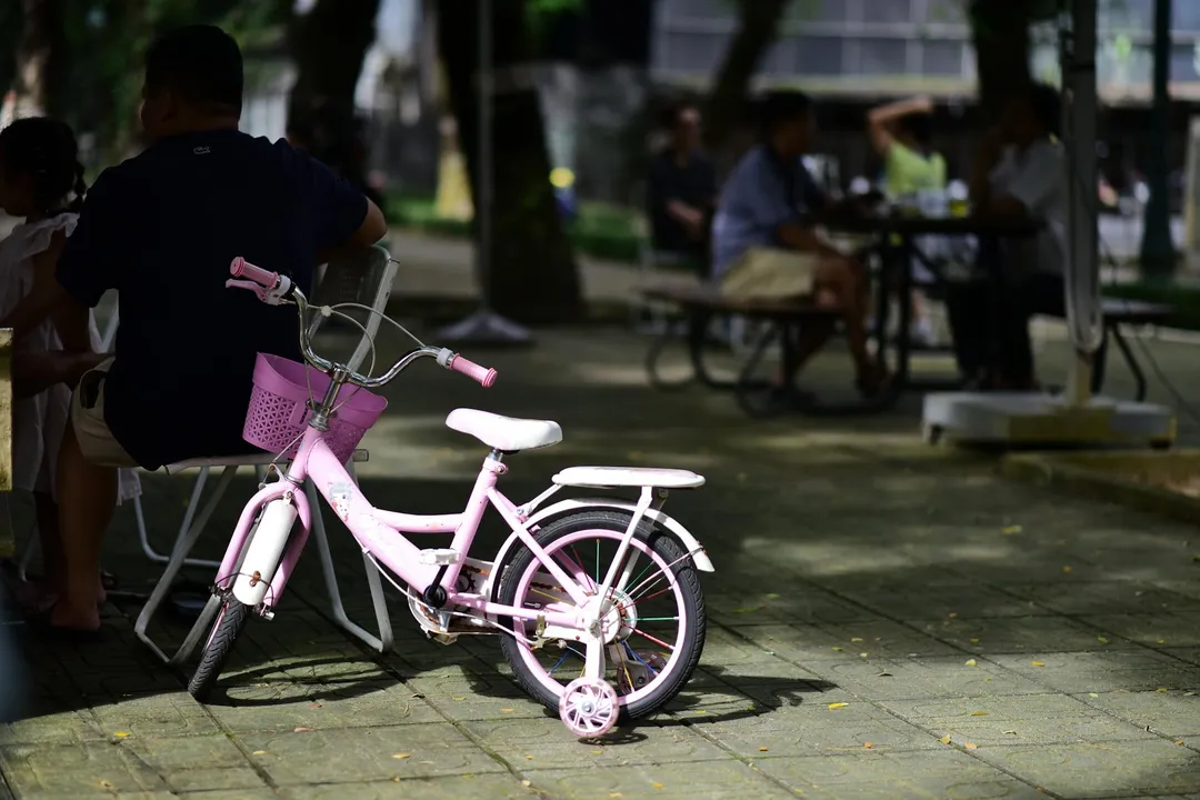 A pink children's bicycle on a paved common-area path with blurred people sitting at tables in the background.