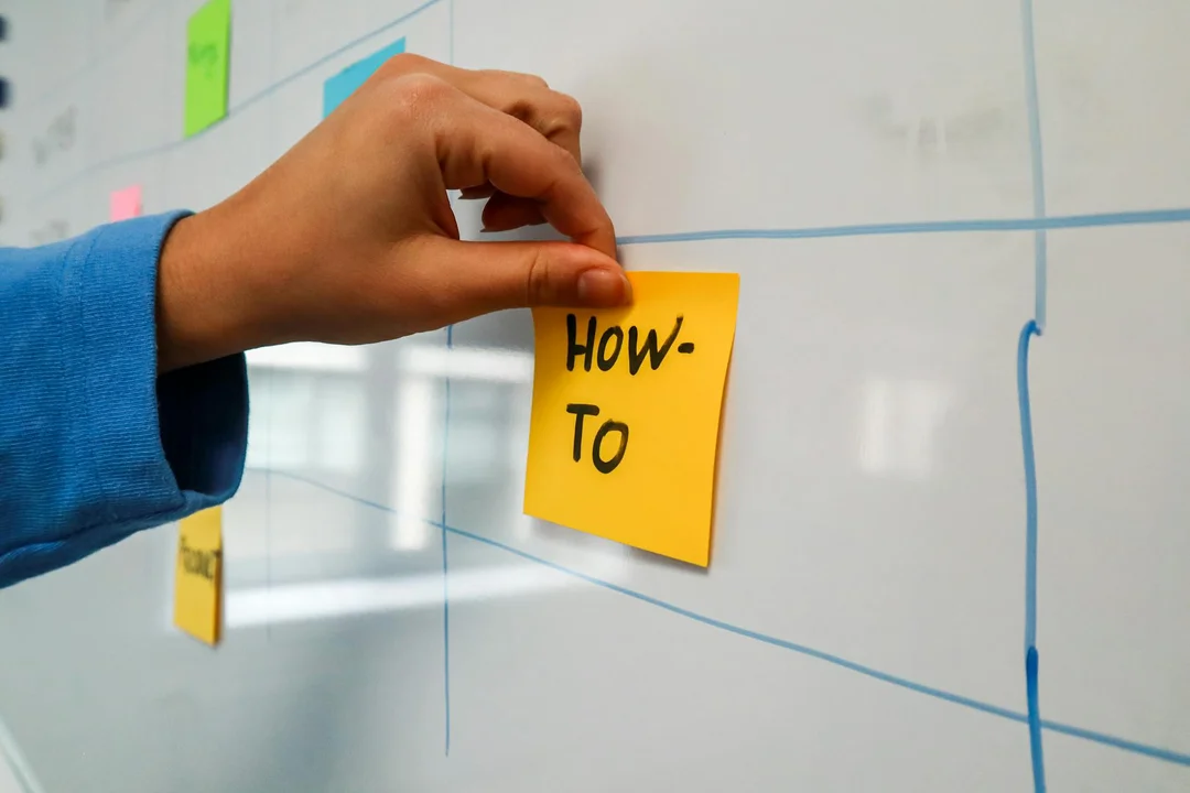 Hand placing a yellow sticky note labeled 'How-To' on a blue planning board with grid lines.