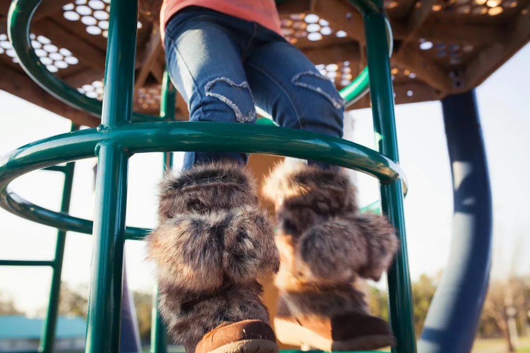 Child climbing a green playground structure, wearing ripped jeans and furry boots.